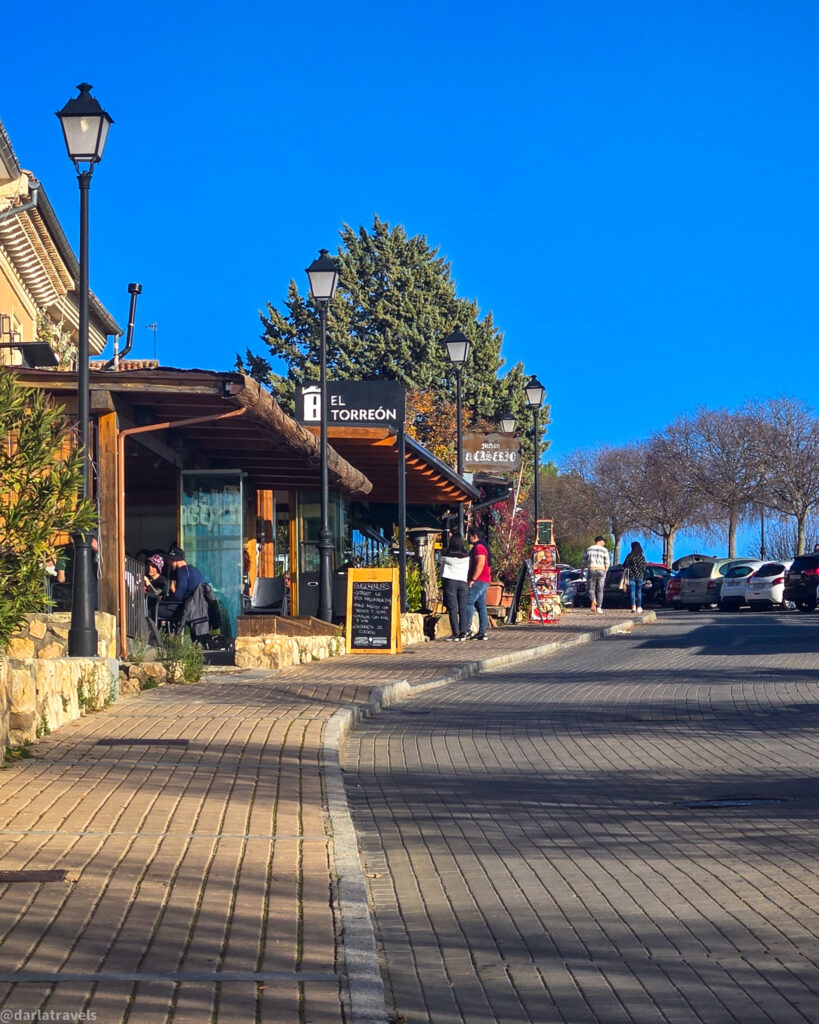 A sunny, eye-level shot of a paved street in Barrio de Castillo, Cuenca, Spain. The street is lined with a sidewalk paved with rectangular stones, and a street with asphalt. On the left, a building with a wooden awning and outdoor seating is visible, with a sign reading "El Torreon." Two people are seated at the outdoor seating. A yellow A-frame sign with a chalkboard is in front of the restaurant. Further down the street, street lamps are visible, along with parked cars and people walking. The sky is a clear, bright blue. 