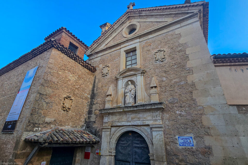 A low-angle, eye-level shot of the Convento de San José de las Carmelitas Descalzas building in Cuenca, Spain. The building has a weathered stone facade with a statue of a figure in a niche above the main entrance. The entrance is a large, dark metal door. A sign to the right of the entrance reads "CONVENTO CARMELITAS" with blue lettering on a white background. A smaller building is attached to the left, with a sign visible on its wall. The sky above is a clear blue.