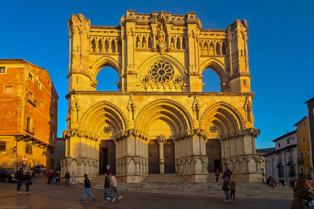  view of the Cuenca Cathedral in Spain. The cathedral is a grand Gothic-style building with an ornately decorated facade featuring numerous sculptures and architectural details. The image highlights the main entrance and the towering spires reaching towards the sky. 