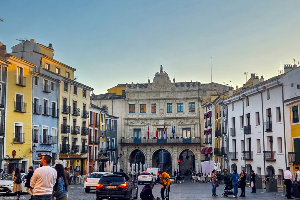 **Description:** The image showcases the Plaza Mayor and town hall in Cuenca, Spain. The town hall is a prominent building with an ornate facade, featuring three arches at the ground level and several flags displayed on the balcony. Surrounding the town hall are colorful buildings with balconies, adding charm to the scene. People are walking and interacting in the plaza, creating a lively atmosphere. Cars are also present, and there are outdoor seating areas with umbrellas, indicating a vibrant and active public space. The image captures the architectural beauty and dynamic energy of this historic location.

Let me know if there's anything else I can assist you with! 😊