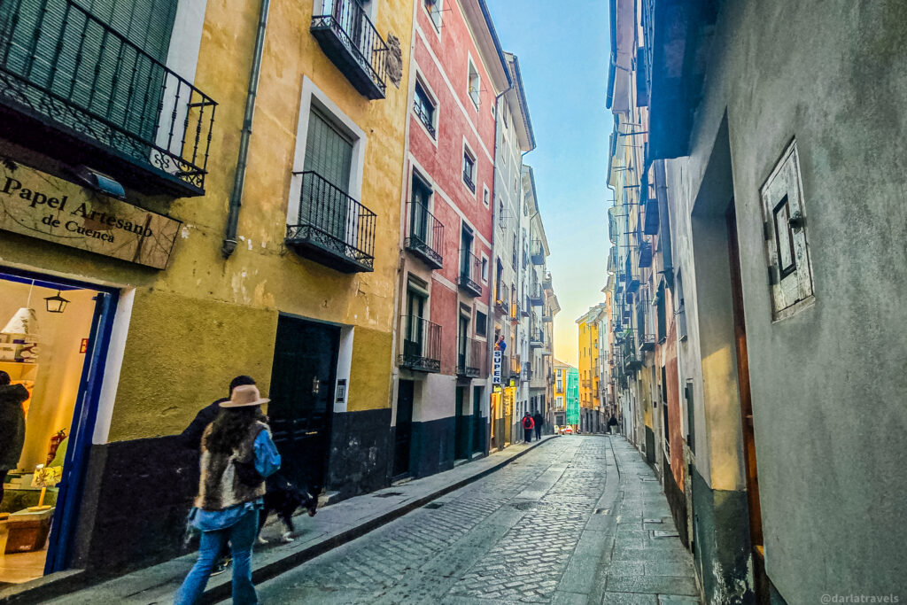 A charming cobblestone street in Cuenca, Spain, lined with colorful, historic buildings with wrought-iron balconies. A woman wearing a hat and fur vest walks her dog past a small artisan paper shop, "Papel Artesano de Cuenca," which has a warmly lit interior. The street narrows into the distance, bathed in soft evening light.