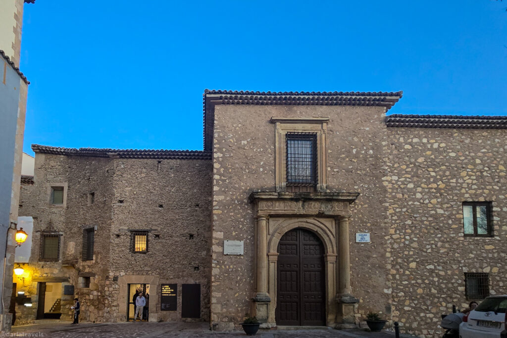 A medium shot of the front of the Hanging Houses in Cuenca, Spain, taken at twilight. The church features a stone facade with a large, arched wooden doorway in the center. A small balcony is situated above the doorway. Warm light emanates from a window on the left side of the building, and a street lamp is visible at the far left. The ground in front of the church is paved with cobblestones. People are partially visible near the entrance. A sign is visible to the right of the door. The sky is a clear, deep blue.