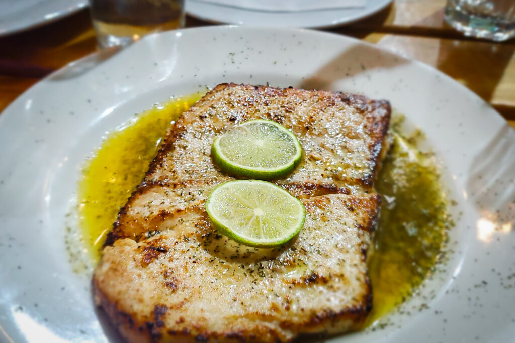 A plated dish featuring two grilled fish fillets, garnished with lime slices and seasoned with herbs and spices. The fillets are accompanied by a rich yellow sauce, likely butter or oil-based, spread elegantly across the plate. This appetizing seafood meal showcases the fresh and flavorful dining options available in Santa Marta, ideal for seafood enthusiasts visiting the area.