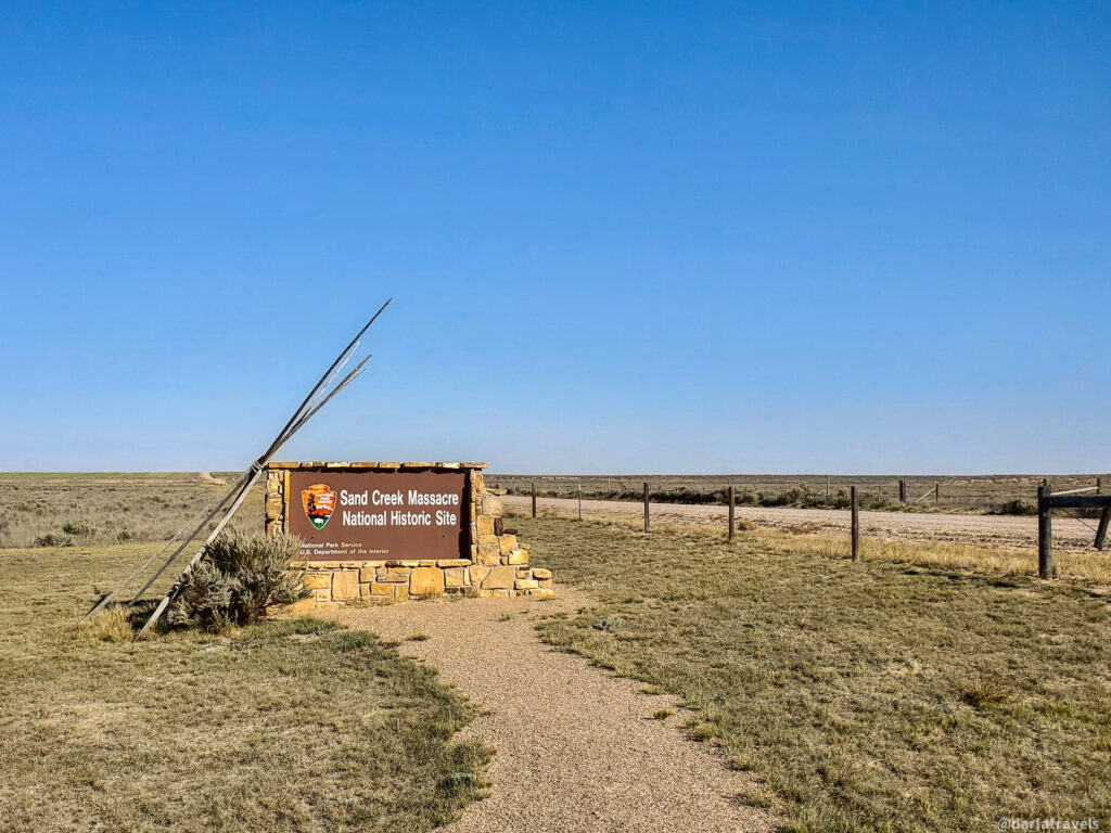 A brown sign that reads "Sand Creek Massacre National Historic Site" stands in a vast, open landscape under a clear blue sky. The sign is supported by two weathered wooden beams that lean against a small, stacked stone structure. In front of the sign, a narrow, winding gravel path leads into the distance. The surrounding terrain is mostly dry, short grassland with a dirt road visible in the midground. A simple wire fence stretches across the landscape in the distance, disappearing towards the horizon. The scene conveys a sense of remoteness and historical significance.