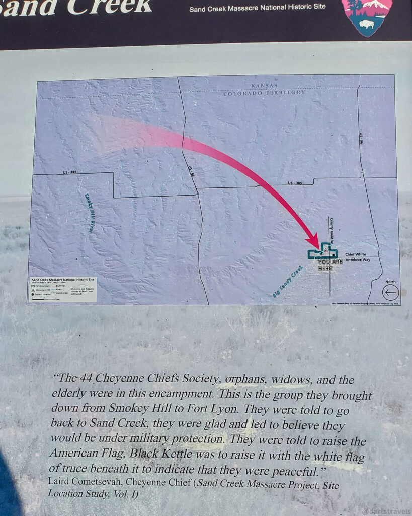 A close-up shot of an informational sign at the Sand Creek Massacre National Historic Site features a map and historical text. The map, located in the upper portion of the sign, shows a portion of the Kansas and Colorado Territory with roads, Sandy Creek, and Big Sandy Creek labeled. A thick pink arrow curves downwards, originating from an area to the northwest and pointing towards a green square labeled "YOU ARE HERE" near Chief White Antelope Way.
The sign also displays the National Park Service logo in the upper right corner and the title "Sand Creek Massacre National Historic Site" at the top. A smaller map with a legend is visible in the lower left corner of the map section.