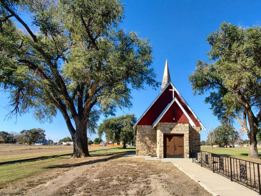 A small, charming chapel, the Kit Carson Memorial Chapel, made of light brown stone with a steep red roof and a white cross on top stands nestled among large green trees under a clear blue sky. The chapel has a pointed steeple and a dark wooden double door at the entrance, flanked by narrow windows. A concrete walkway leads up to the chapel, bordered by a low, decorative black metal fence on the right. The surrounding area features a mix of dry, grassy fields and green lawns, with scattered trees providing shade. In the distance, hints of houses and a road can be seen, suggesting a peaceful, rural or suburban setting. The sunlight casts shadows of the trees and the chapel onto the ground.