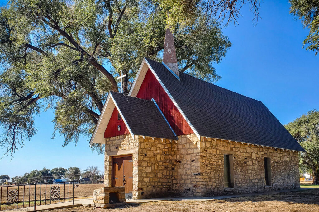 The Kit Carson Memorial Chapel at Fort Lyon National Cemetery. A small, rustic chapel constructed from light brown stone with a dark shingle roof stands under a clear blue sky, partially shaded by large trees with green leaves. The chapel features a steep, red-trimmed gable roof with a simple white cross mounted on the front. A dark wooden door serves as the entrance, and narrow rectangular windows are visible on the side. A low, black metal fence encloses a small area in front of the chapel. The ground surrounding the building is a mix of dry grass and dirt. In the blurred background, a flat, open field with sparse vegetation and distant houses can be seen. The sunlight casts shadows from the trees onto the chapel and the ground.