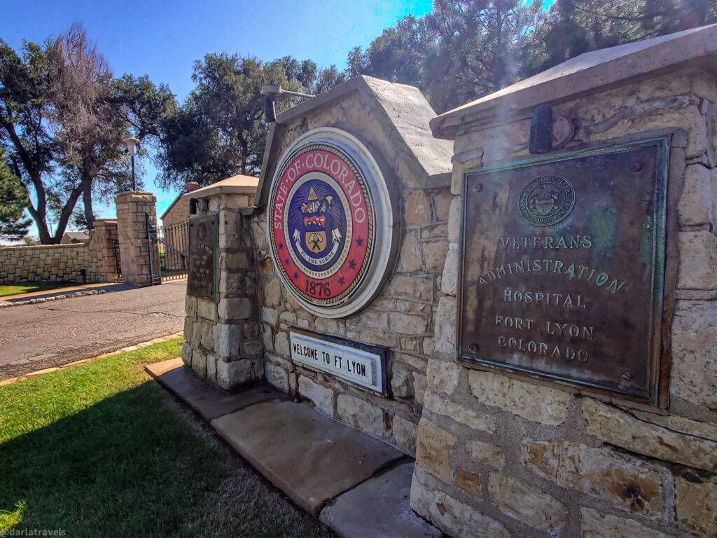 A stone entrance gate under a bright sky features the Seal of the State of Colorado prominently displayed on the left pillar. The circular seal has a colorful design with the words "STATE OF COLORADO" around the top and "1876" at the bottom. Below the seal, a smaller white sign reads "WELCOME TO FT LYON."
On the right stone pillar, a weathered bronze plaque is affixed, reading "VETERANS ADMINISTRATION HOSPITAL FORT LYON COLORADO." The stone of the pillars is light brown and textured, showing signs of age and weathering.
Beyond the gate, a glimpse of green grass, trees, and a brick building with a fence can be seen. The sunlight casts shadows on the stone pillars and the surrounding area. The scene suggests the entrance to a historical or significant site associated with the State of Colorado and a Veterans Administration hospital.