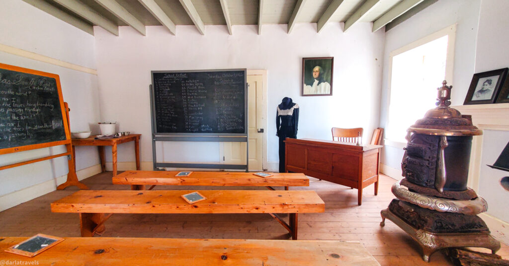 Alt text generator
Custom Gem
A brightly lit, historical one-room schoolhouse with wooden floors and white walls. Two long, light-colored wooden desks with benches face two black chalkboards on the left wall. On the first chalkboard, white chalk writing is partially visible. The second chalkboard displays a neatly written list of names and numbers in white chalk. A small wooden table with a white bowl and other items sits to the left of the second chalkboard.
In the back of the room, a wooden teacher's desk and chair are positioned in front of a window that lets in bright daylight. A framed portrait of a man hangs on the wall above the desk. To the right, a vintage, ornate wood-burning stove stands on the wooden floor. Another framed picture is visible on the wall above the stove. A doorway in the back of the room shows a coat hanging on a hook. The ceiling has exposed wooden beams painted white.