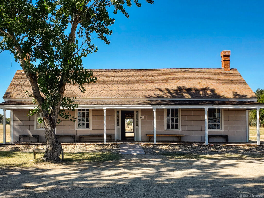 The Prowers House at Boggsville Historic Site. A long, single-story building with a light brown exterior and a shingle roof stands under a clear blue sky. A covered porch supported by white columns runs along the front of the building, providing shade. Several evenly spaced windows with white frames line the front facade. A brick chimney rises from the roof on the right side. A dark doorway is located centrally under the porch. Wooden benches are placed along the porch. A large tree with green leaves stands to the left of the building, casting shadows on the ground. The ground in front of the building is a mix of dirt and sparse grass. The overall scene suggests a historic building in a rural or open setting.