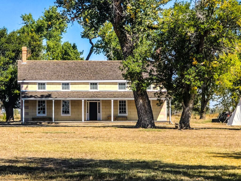 This is the Prowers House at Boggsville Historic Site. A two-story, light yellow house with a brown shingle roof stands in a grassy field dotted with large, leafy trees under a clear blue sky. The house has a covered porch supported by white columns running along the front of the first floor, with several evenly spaced windows on both the ground and upper levels. A brick chimney rises from the left side of the roof. A dark doorway is located in the center of the first floor, leading onto the porch. The surrounding field is dry and golden, suggesting late summer or early autumn. The large trees cast shadows on the lawn. In the far right, a glimpse of a white tent-like structure can be seen.