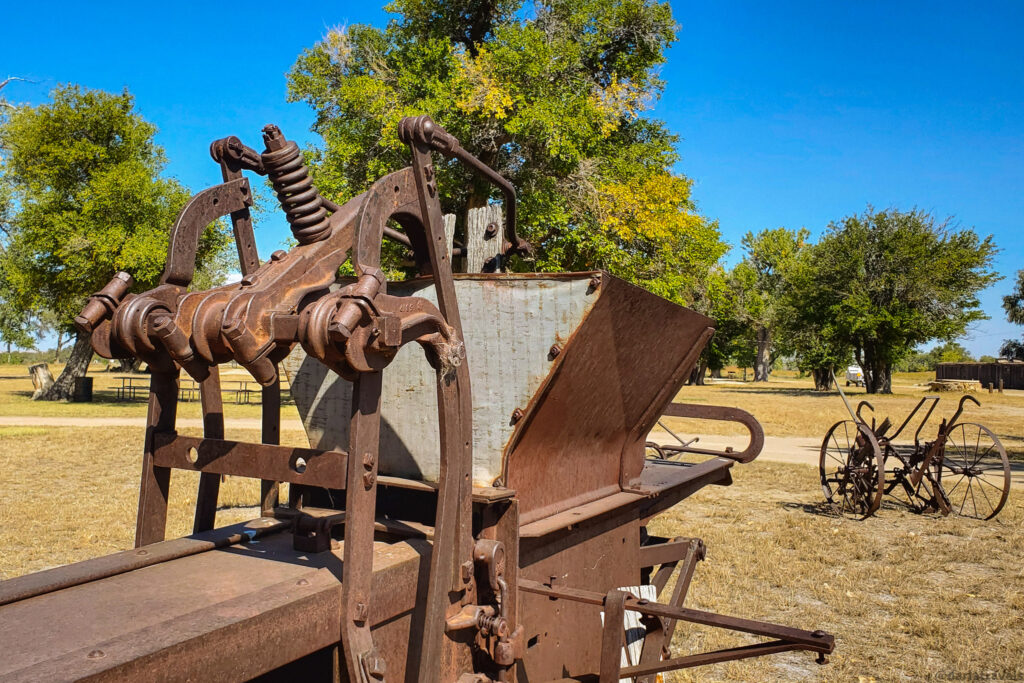 A rusty, antique farm machine dominates the foreground, set against a backdrop of a grassy field, scattered trees with green and yellow leaves, and a clear blue sky. The machine is complex, with various metal parts, gears, springs, and a hopper-like structure. Its weathered appearance suggests it has been sitting outdoors for a long time. In the middle ground, another smaller, rusty piece of farm equipment with large wheels is visible. The background features more trees and a hint of a wooden fence or structure in the distance. The bright sunlight casts shadows on the ground, highlighting the textures of the old machinery and the natural landscape.