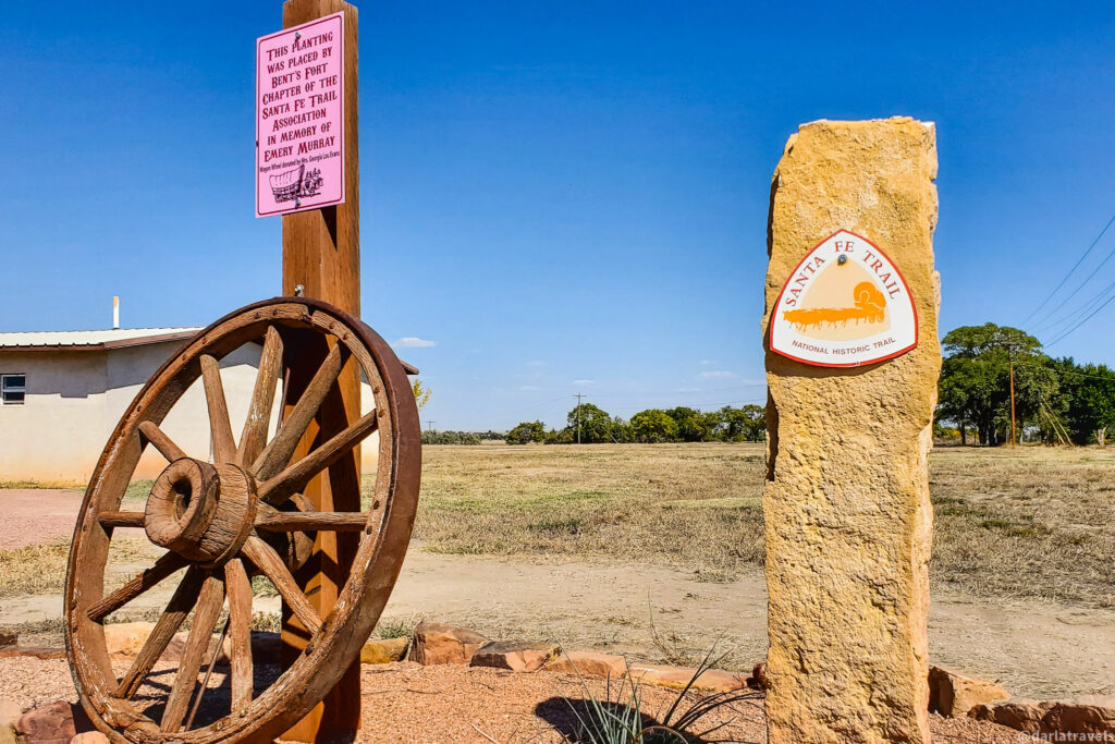A weathered wooden wagon wheel leans against a wooden post with a pink sign in a sunny, rural setting. The large, brown, spoked wheel rests on reddish-brown gravel. The pink sign attached to the post above the wheel has black lettering. To the right, a light brown, textured stone marker stands upright, featuring a circular sign that reads "SANTA FE TRAIL" in red letters around a white center, with "NATIONAL HISTORIC TRAIL" written below in smaller red letters. The background shows a dry, grassy field under a clear blue sky, with sparse trees visible in the distance. A white, low building with a flat roof is partially visible on the left side of the image.