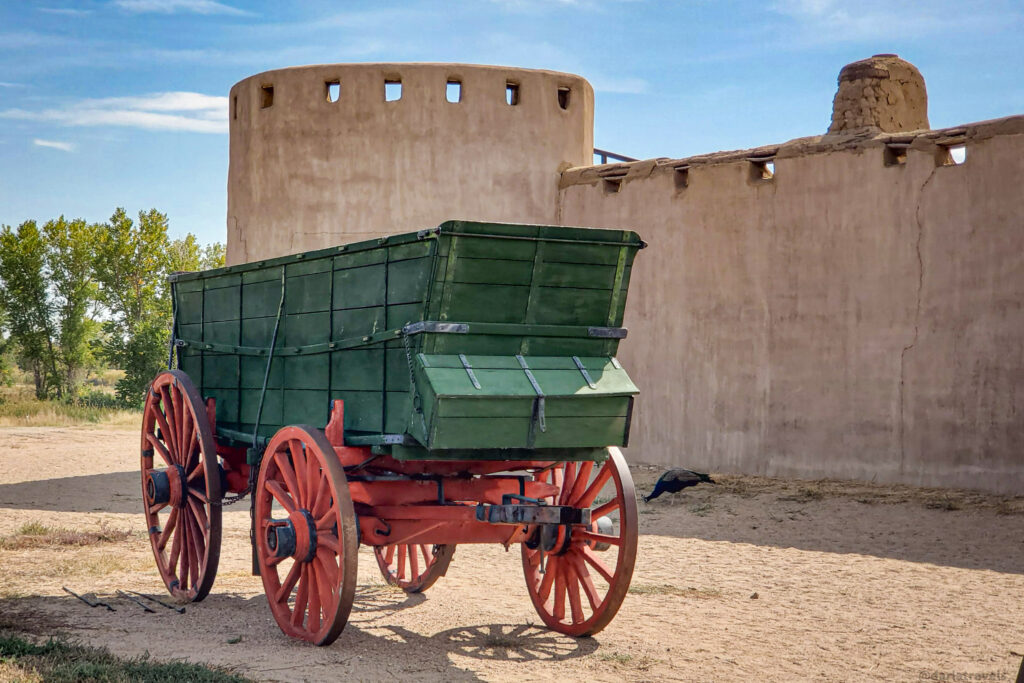 A green and red wooden wagon sits on a dirt ground in front of a light brown adobe building under a blue sky with scattered clouds. The wagon has large red spoked wheels and a green boxy carriage. The adobe building features a round tower-like structure on the left with small rectangular openings near the top and a longer, lower section to the right with similar openings. A dark bird walks on the ground near the wagon. In the background, green trees are visible on the left.