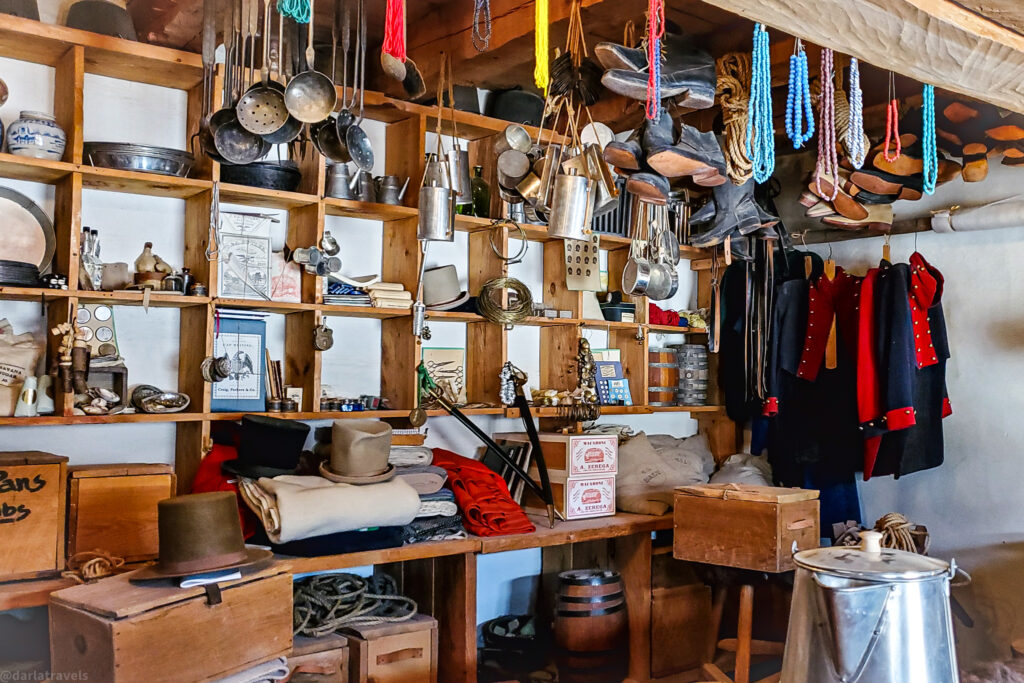 The interior of a room at Bent's Old Fort is filled with a diverse collection of historical objects, meticulously arranged on wooden shelves and hanging from the ceiling and walls. On the left side, shelves hold various items, including ceramic jars, metal bowls, papers, shoes, and small figurines. Below these shelves, wooden crates and rolled-up textiles are stacked.
Above, numerous metal pots, pans, and utensils hang from hooks attached to the ceiling. In the center of the room, more shelves display hats, tools, boxes, and a sword. To the right, boots, moccasins, and beaded necklaces dangle from above, while several red and blue military-style jackets hang against the far wall. A small wooden barrel sits on a lower shelf, and a large metal pot with a lid rests on the floor in the right foreground. The room has a rustic feel, illuminated by natural light filtering in from an unseen source, highlighting the textures and variety of the historical artifacts. This scene offers a glimpse into the daily life and supplies of Bent's Old Fort..