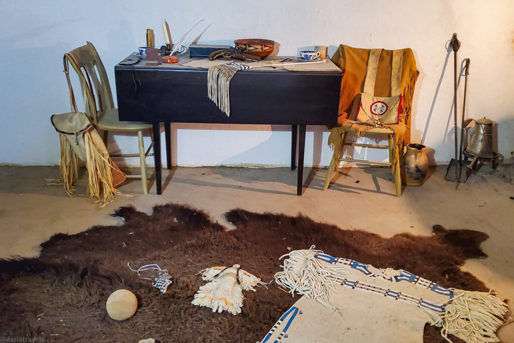 A dimly lit room features a collection of historical artifacts arranged to depict a scene from the past within Bent's Old Fort. A dark wooden drop-leaf table stands against a white wall, adorned with various items including quill pens, inkwells, a small beaded bag, a pipe, and a wooden bowl. A fringed garment or textile is draped over the edge of the table.
Two simple wooden chairs are positioned at the table. The chair on the left has a fringed pouch or bag hanging from its back. The chair on the right is draped with a light brown garment and has a decorated pouch resting on it.
On the floor, a large brown fur hide dominates the foreground. Several smaller items are scattered on the hide, including a smooth, light-colored stone, a pair of beaded moccasins, and a partially visible beaded and fringed textile.
In the background, against the right wall, stand a few metal implements, including what appears to be a branding iron and a metal container. A simple clay pot rests on the floor near the chair on the right. The overall ambiance suggests a re-creation of a living or working space from the 19th century fur trade era.