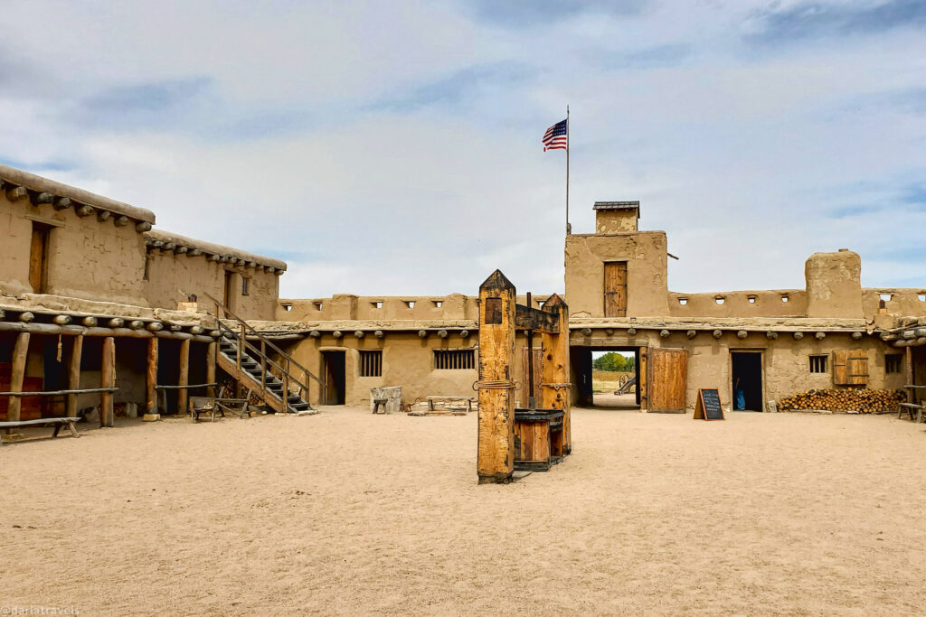 The interior courtyard of Bent's Old Fort, featuring adobe buildings with wooden supports, a central wooden structure, and the American flag flying atop a tower under a partly cloudy sky.