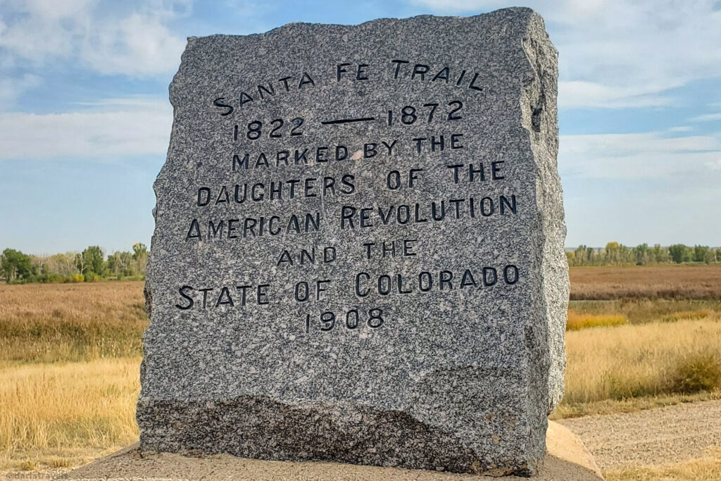 A weathered gray stone monument stands in a field of dry grasses under a partly cloudy sky. Inscribed in bold, sans-serif lettering are the words "SANTA FE TRAIL," followed by "1822 - 1872." Below this, the inscription continues, "MARKED BY THE DAUGHTERS OF THE AMERICAN REVOLUTION AND THE STATE OF COLORADO 1908." The monument rests on a slightly lighter-colored stone base. In the blurred background, a line of trees marks the horizon.