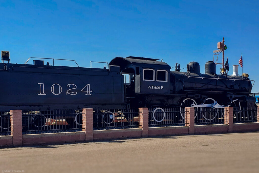 A vintage black AT&SF steam locomotive, #1024, is displayed outdoors behind a fence under a clear blue sky. On display in La Junta, Colorado rail park.