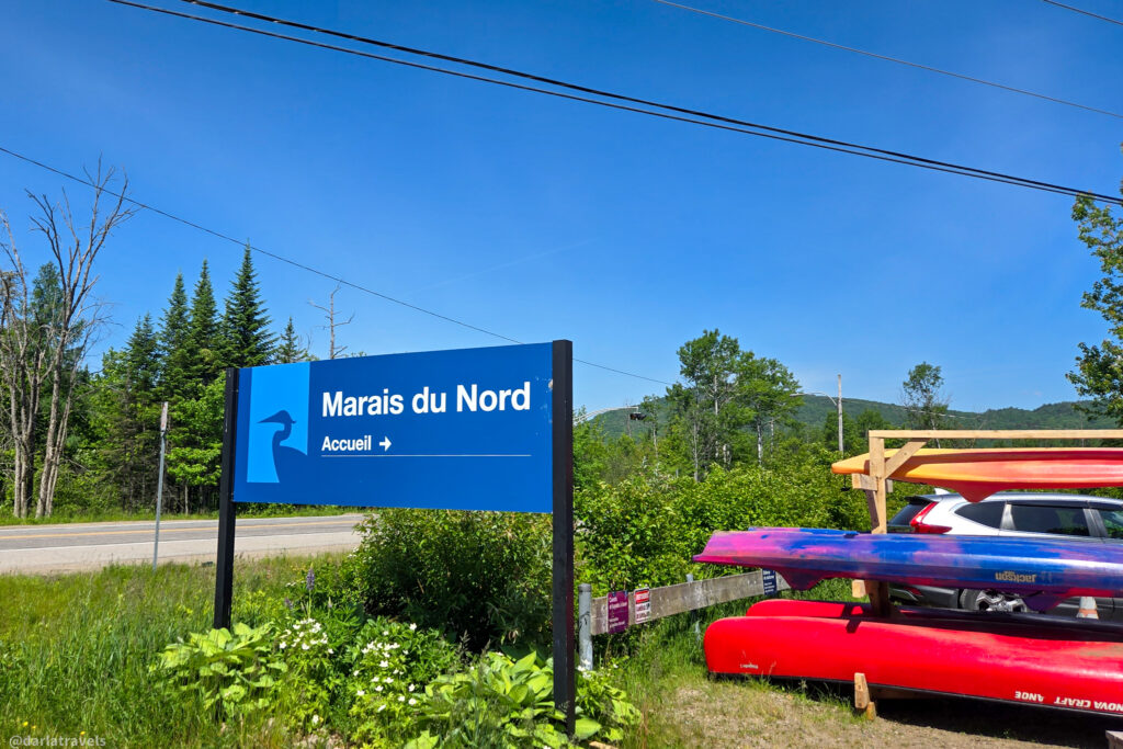 Blue directional sign for Marais du Nord beside parked car and stacked kayaks in wooded natural area.