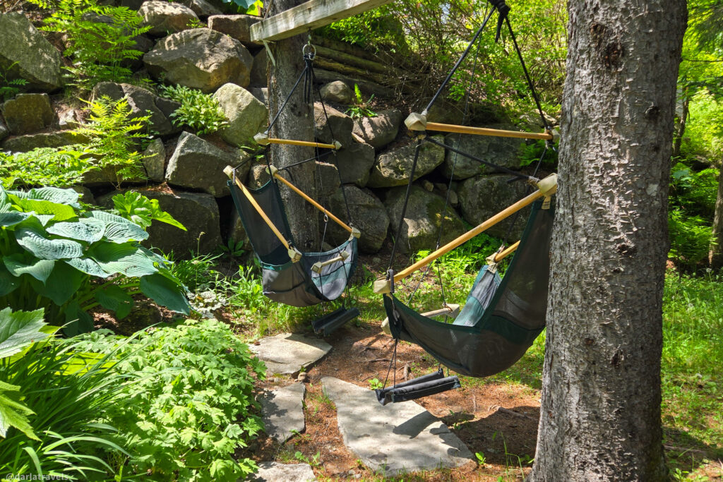 Two hanging chairs suspended from wooden beam in forest setting, with stone wall, greenery, and stone pathway.