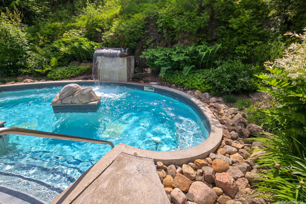 Small stone-edged hot tub pool with waterfall feature, surrounded by greenery and metal handrail for entry.