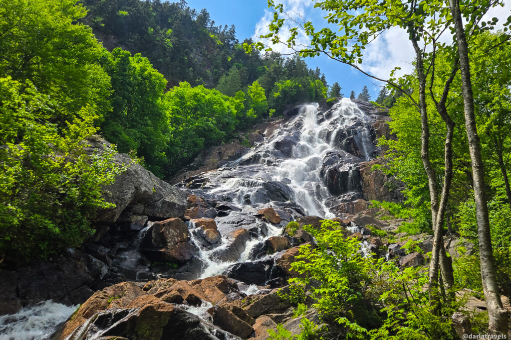 Chute Delaney waterfall cascading down dark rocks amid trees and blue sky in Vallée Bras-du-Nord