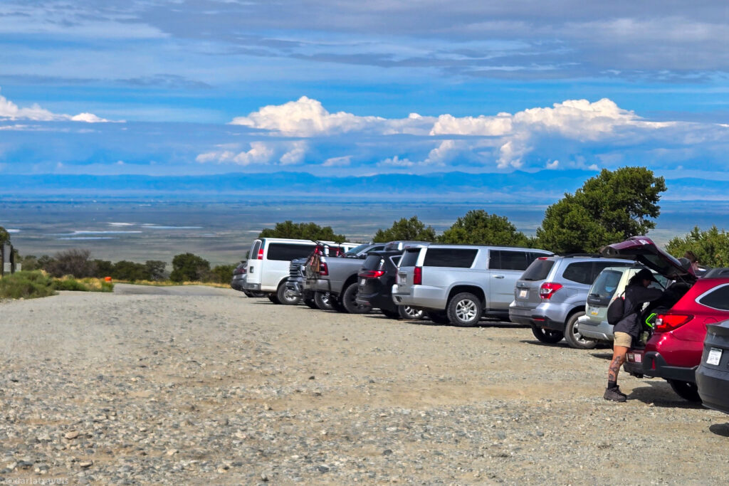 Gravel parking area near Zapata Falls trailhead overlooking the San Luis Valley with San Juan mountains in the distance. Parked vehicles and hiker preparing gear under sunny sky.