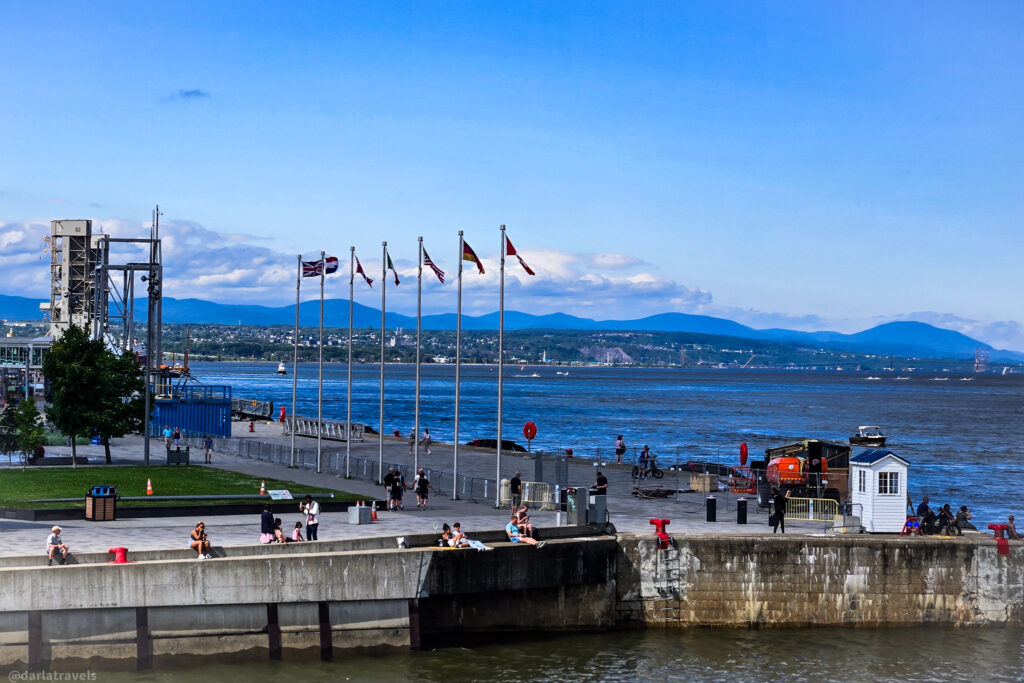 Waterfront promenade with flags, red chairs, and scenic view across river in Quebec City. Laurentian mountains in the background