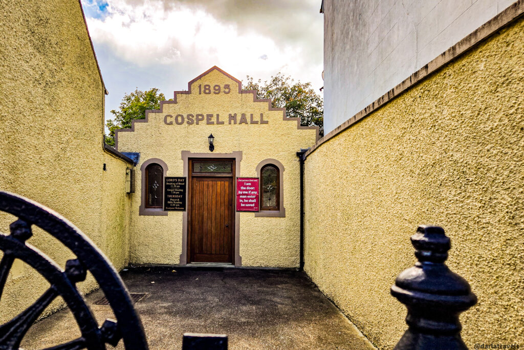 Narrow entrance to a yellow-painted Gospel Hall with a distinctive stepped gable marked 1895, wooden door, and sign boards.