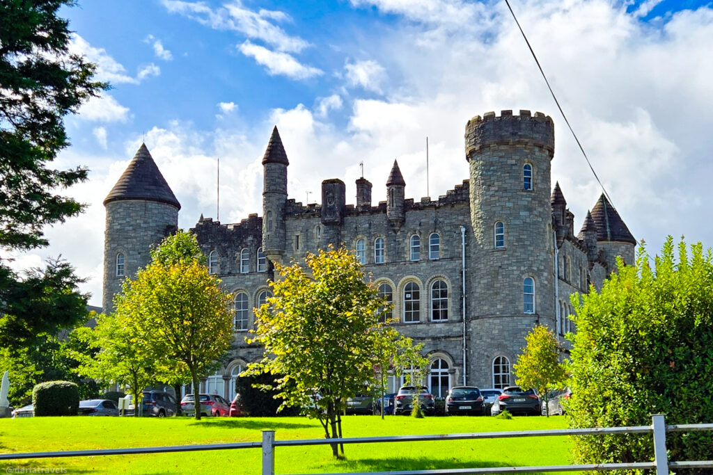 A stone castle-like building with towers and arched windows stands behind a green lawn, under a blue sky with scattered clouds.
