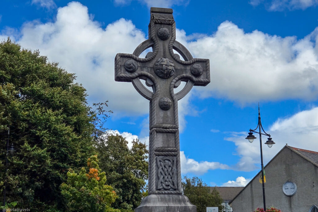 A large Celtic cross memorial with detailed carvings rises against a bright blue sky, surrounded by trees and buildings. Letterkenny, County Donegal, Ireland