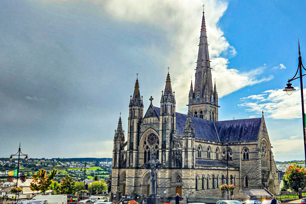 Large Gothic-style church with tall spires and detailed stonework, overlooking a valley of houses and green fields.