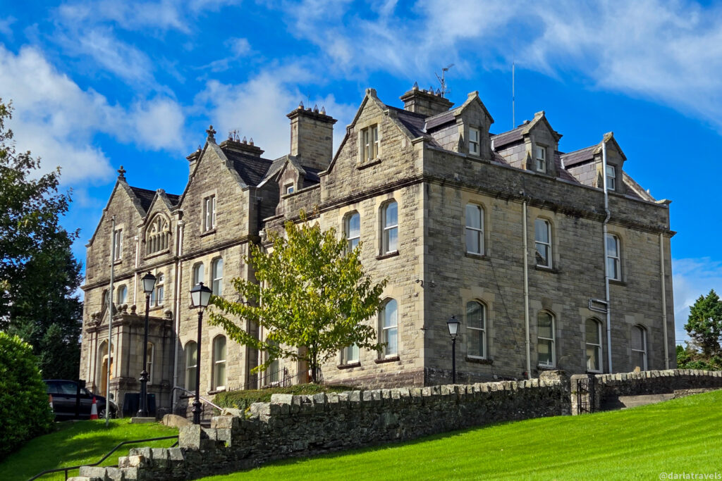 Historic stone building with arched windows and a pitched roof, set on a green lawn beneath a bright blue sky. Viewable on a walking tour of Letterkenny, County Donegal.