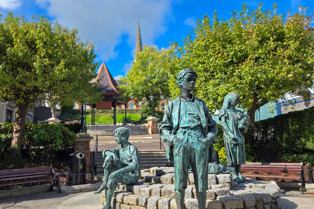 Bronze statues of children in a public square surrounded by trees and benches, with church spire visible in the background; stop on a walking tour of Letterkenny, County Donegal, Ireland