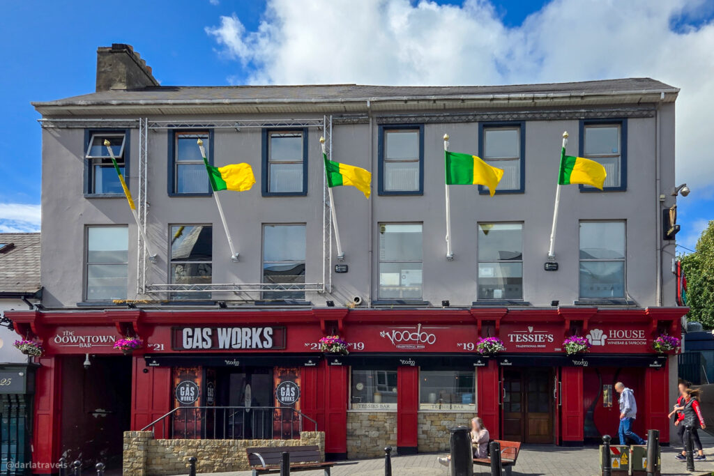 Traditional pub and shops with red storefronts, hanging flower baskets, and green-yellow flags above, while people walk past.