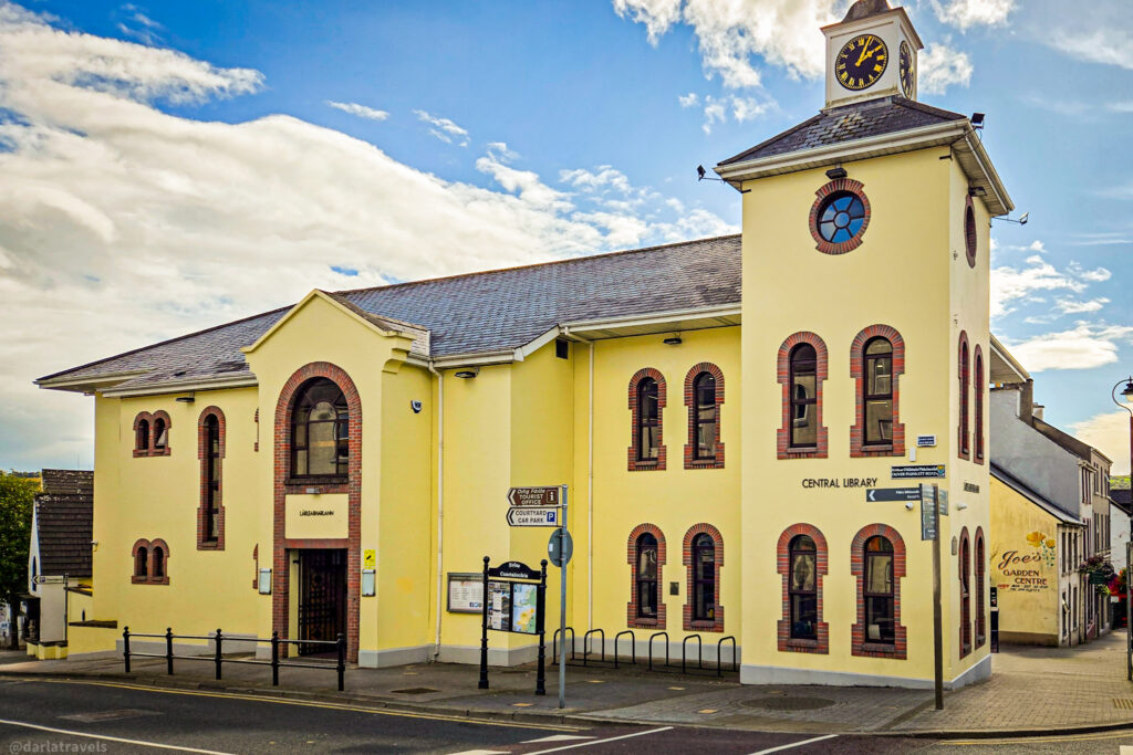 Yellow-painted central library building with arched windows and a clock tower, set on a street corner under a bright blue sky.