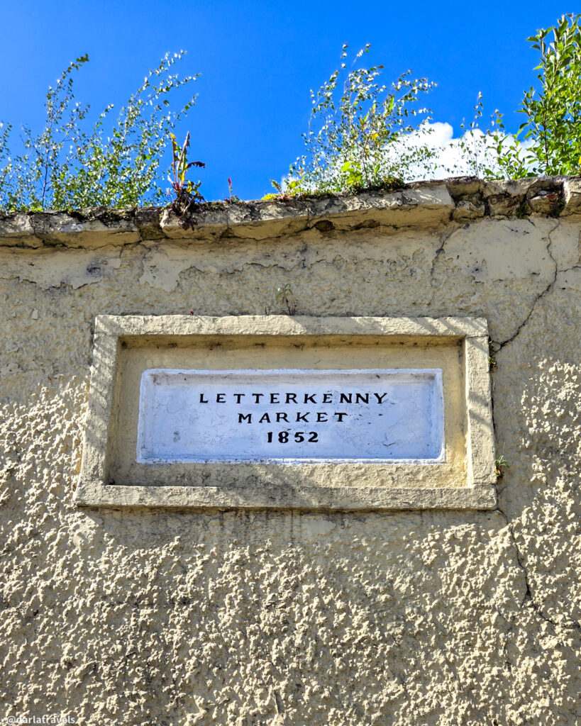 Historic stone plaque reading "Letterkenny Market 1852" is set into a textured wall, with plants growing on top against a blue sky.