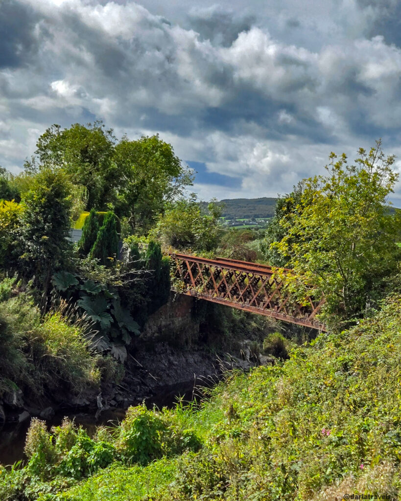Iron railway bridge crosses a lush green ravine with trees and shrubs, set against a dramatic sky and distant rolling hills.