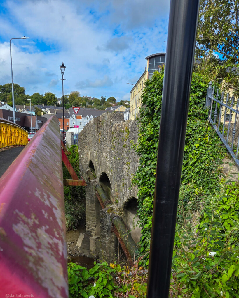 Stone arch bridge with ivy-covered walls and pipes running beneath, located beside city buildings and greenery under a partly cloudy sky.