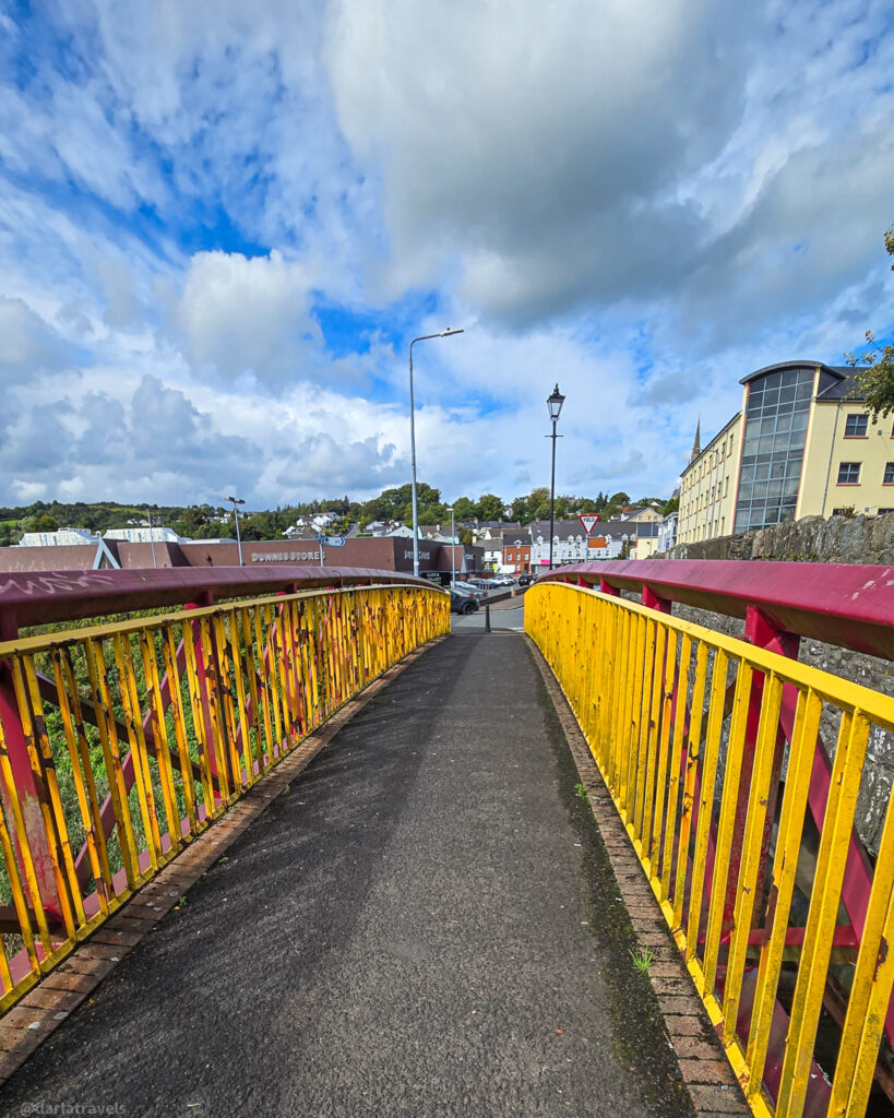 Yellow and red pedestrian bridge with metal railings leads into town, with shops and lamp posts visible ahead under wide-clouded skies.