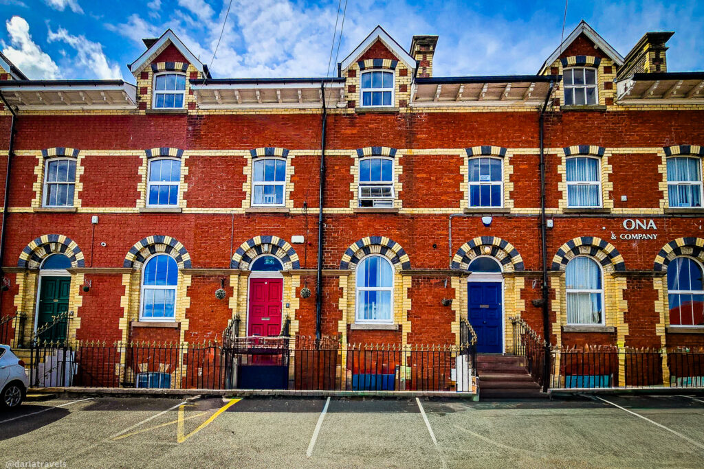 Victorian red-brick row houses with patterned arches, colored doors, and parking spaces in front under a partly cloudy sky.