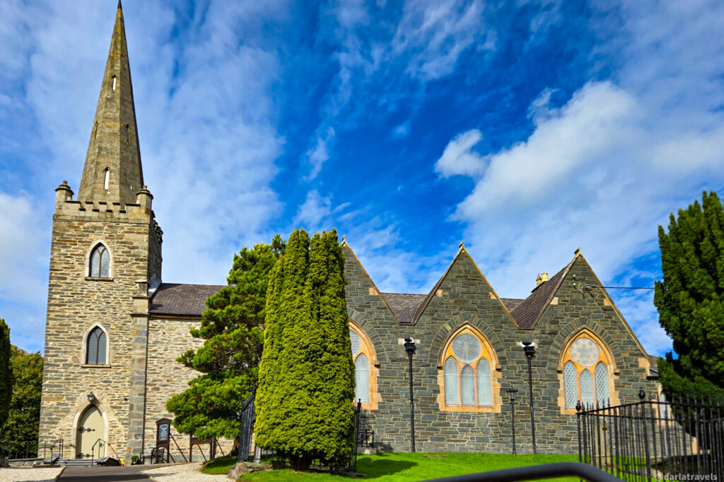 Stone church with tall steeple and arched windows, flanked by manicured trees and shrubs, sitting beneath a vibrant blue sky.