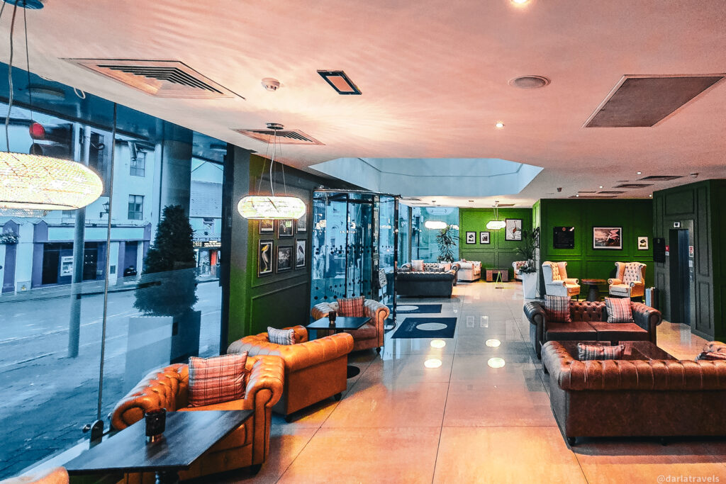 Modern hotel lounge with brown leather sofas, plaid cushions, green walls, and large windows overlooking a city street.
