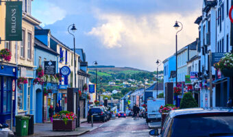 Colorful main street with shops and flower boxes, cars parked along the road, and rolling green hills visible in the background.