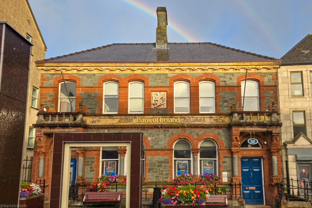 Colorful main street with shops and flower boxes, cars parked along the road, and rolling green hills visible in the background.