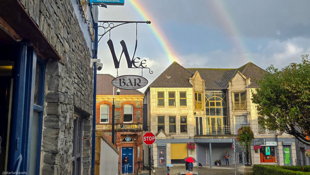 Stone building with a metal “Wee Bar” sign, rainbow in the sky, and people walking in a lively town square with shops nearby.