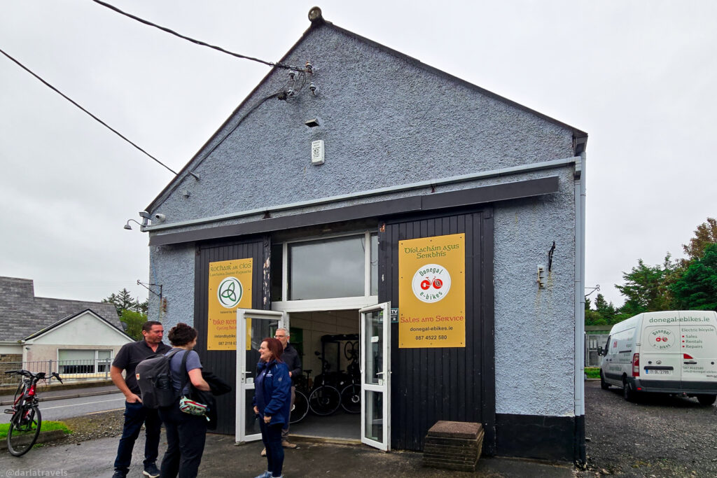 Four people stand outside a grey, textured building advertising Donegal e-bikes with yellow signage on a cloudy day.
