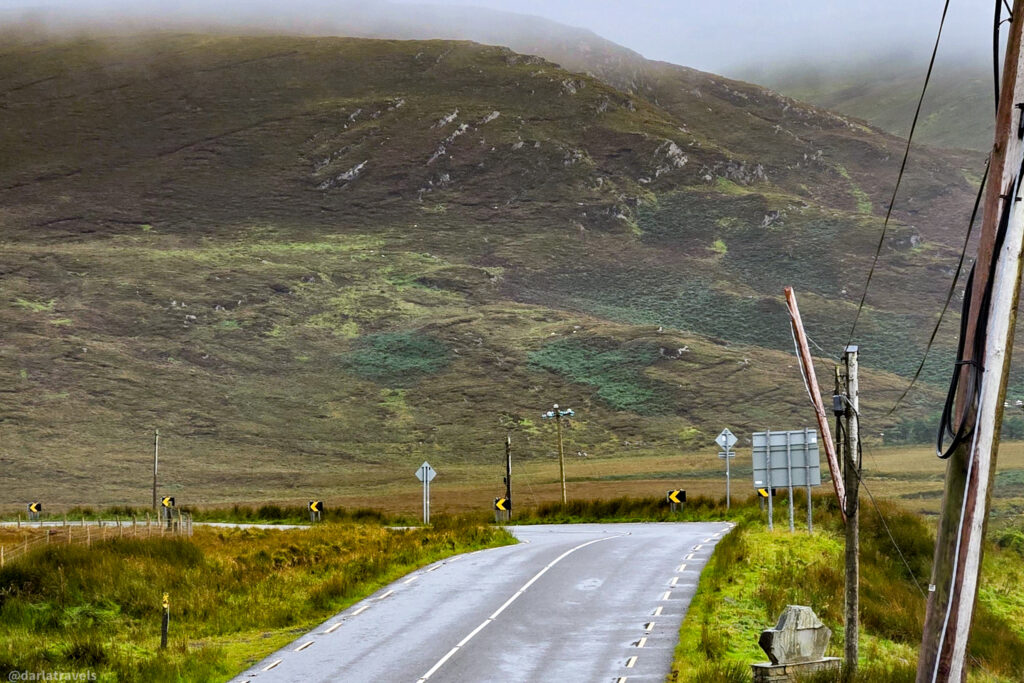 Wet asphalt road curves past grassy verges and utility poles toward a large, fog-shrouded, moss-covered mountain.