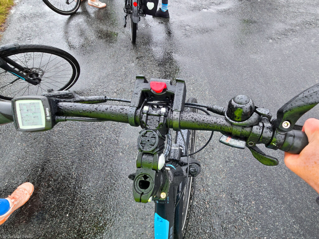 First-person view of wet black bicycle handlebars featuring a Bosch display, bell, and hand gripping the wet pavement surface with other cyclists nearby.